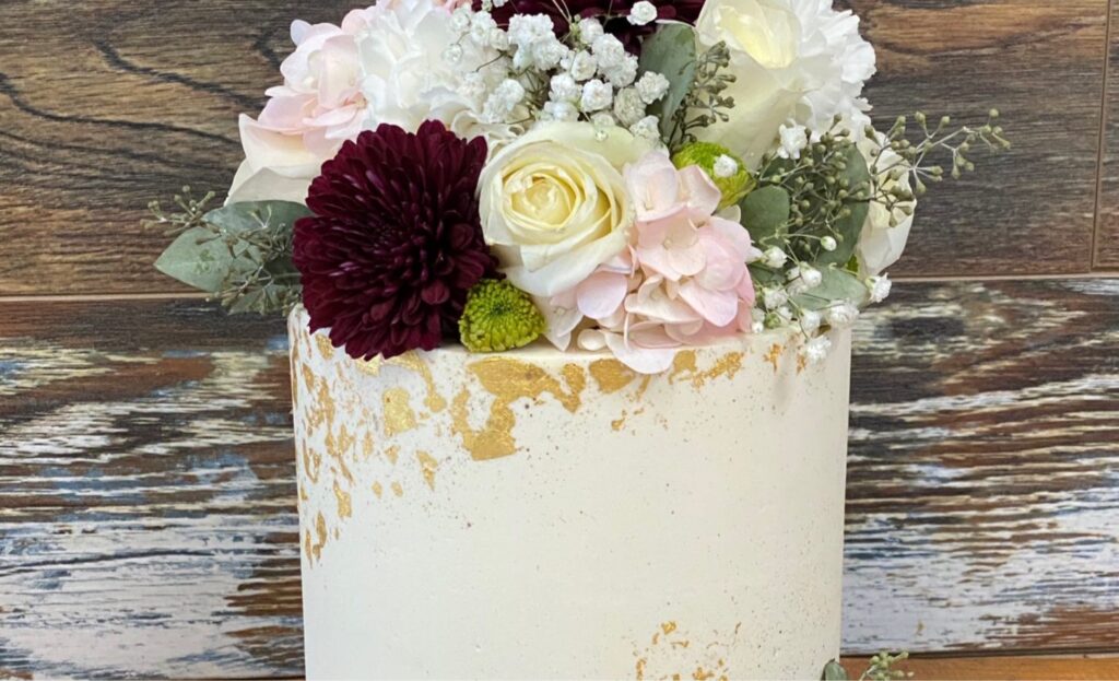 White celebration cake with gold leaf and fresh flowers on a rustic wooden table