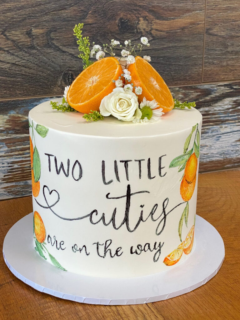 Baby shower cake hand lettered "Two little cuties are on the way" with oranges and flowers